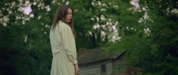 Movie still from “Still” (2018), directed by Takashi Doscher – A woman in a white dress standing in front of trees; Medium shot, Low angle