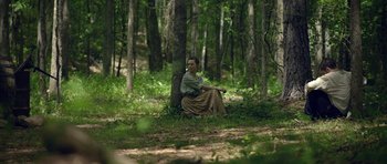 Movie still from “Still” (2018), directed by Takashi Doscher – A woman sitting on the ground next to a tree in the woods; Wide shot, High angle