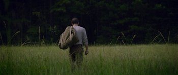 Movie still from “Still” (2018), directed by Takashi Doscher – A man walking through tall grass holding a bag; Wide shot, Low angle