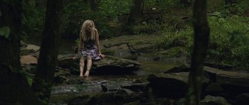 Movie still from “Still” (2018), directed by Takashi Doscher – A woman sitting on a rock near a stream; Wide shot, High angle