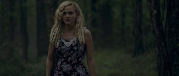 Movie still from “Still” (2018), directed by Takashi Doscher – A woman with blonde dreadlocks standing in the woods; Medium shot, Over the shoulder angle