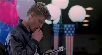 Movie still from “Stormy Monday” (1988), directed by Mike Figgis – A young man reading a newspaper in front of american flags; Medium shot, Over the shoulder angle