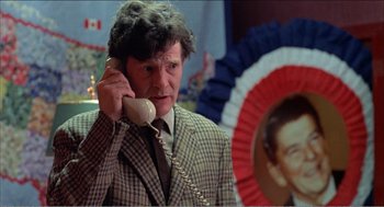 Movie still from “Stormy Monday” (1988), directed by Mike Figgis – A man talking on a phone while standing next to an image of ronald reagan; Close Up shot, High angle