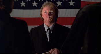 Movie still from “Stormy Monday” (1988), directed by Mike Figgis – A man wearing a suit and tie standing in front of an american flag; Close Up shot, Low angle
