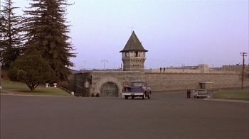 Movie still from “Straight Time” (1978), directed by Ulu Grosbard – A car parked in front of a stone wall with a clock tower in the background; Extreme Wide shot, High angle