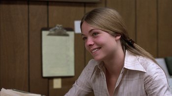 Movie still from “Straight Time” (1978), directed by Ulu Grosbard – A woman sitting at a desk smiling for the camera; Close Up shot, Over the shoulder angle