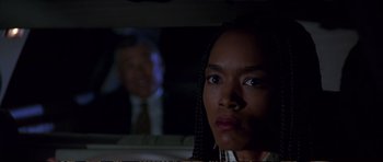 Movie still from “Strange Days” (1995), directed by Kathryn Bigelow – A woman in a car looking at the camera with a man in the background; Close Up shot, Over the shoulder angle