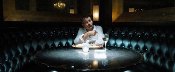 Movie still from “Stretch” (2014), directed by Joe Carnahan – A man sitting at a table with a plate of food and a glass of wine in front of him; Medium shot, High angle