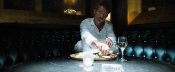 Movie still from “Stretch” (2014), directed by Joe Carnahan – A man sitting in front of a plate of food on top of a table; Medium shot, Over the shoulder angle