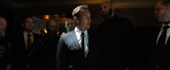 Movie still from “Stretch” (2014), directed by Joe Carnahan – A man in a suit and tie standing next to a group of men; Medium shot, Low angle