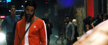 Movie still from “Stretch” (2014), directed by Joe Carnahan – A man in a red and white track suit standing in front of a group of people; Medium shot, Over the shoulder angle