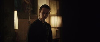 Movie still from “Suburra” (2015), directed by Stefano Sollima – A man in a priest's outfit standing in front of a lamp; Medium shot, Over the shoulder angle