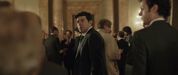 Movie still from “Suburra” (2015), directed by Stefano Sollima – A man in a suit standing in front of a group of people; Medium shot, Over the shoulder angle