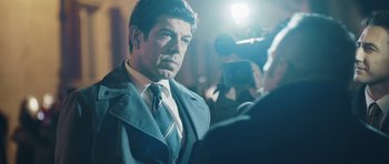 Movie still from “Suburra” (2015), directed by Stefano Sollima – A man wearing a suit and tie while being interviewed by a camera; Medium shot, Over the shoulder angle
