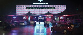 Movie still from “Suburra” (2015), directed by Stefano Sollima – A building that has lights on it and people walking around; Extreme Wide shot, Low angle