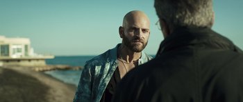 Movie still from “Suburra” (2015), directed by Stefano Sollima – A man with a bald head talking to another man on the beach; Close Up shot, Over the shoulder angle