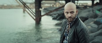 Movie still from “Suburra” (2015), directed by Stefano Sollima – A man with a bald head standing in front of a body of water; Close Up shot, Over the shoulder angle