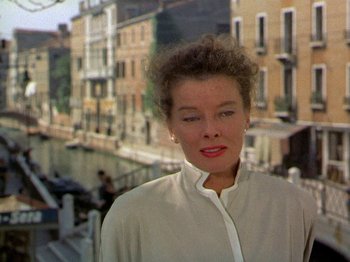 Movie still from “Summertime” (1955), directed by David Lean – A woman in a white shirt standing in front of some buildings; Close Up shot, Low angle