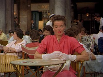 Movie still from “Summertime” (1955), directed by David Lean – A woman sitting at an outdoor table with her hands in her gloves; Medium shot, Over the shoulder angle