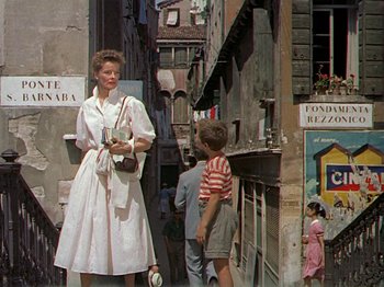 Movie still from “Summertime” (1955), directed by David Lean – A woman in a white dress standing next to a boy in a striped shirt; Wide shot, Low angle