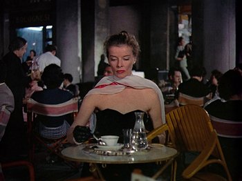 Movie still from “Summertime” (1955), directed by David Lean – A woman sitting at a table with a cup of coffee; Medium shot, Over the shoulder angle