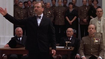 Movie still from “Sunshine” (1999), directed by István Szabó – A man in a suit and tie standing in front of a group of people; Medium shot, Low angle