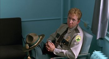 Movie still from “Super Troopers” (2001), directed by Jay Chandrasekhar – A man sitting on a chair wearing a hat; Medium shot, Low angle