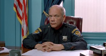 Movie still from “Super Troopers” (2001), directed by Jay Chandrasekhar – A police officer sitting at a desk in front of papers; Close Up shot, Low angle