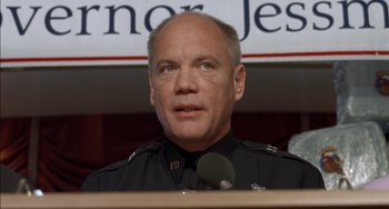 Movie still from “Super Troopers” (2001), directed by Jay Chandrasekhar – A man in a police uniform is sitting at a podium; Close Up shot, Low angle