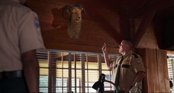 Movie still from “Super Troopers 2” (2018), directed by Jay Chandrasekhar – A man standing next to a ram's head in a room; Medium shot, Over the shoulder angle