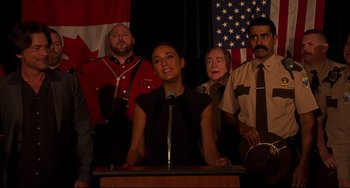 Movie still from “Super Troopers 2” (2018), directed by Jay Chandrasekhar – A woman standing at a podium in front of a group of people; Medium shot, Over the shoulder angle