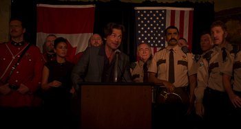 Movie still from “Super Troopers 2” (2018), directed by Jay Chandrasekhar – A man standing at a podium in front of a group of people; Medium shot, Over the shoulder angle
