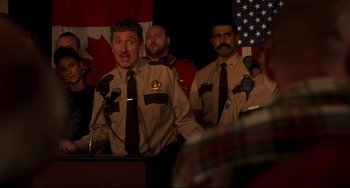 Movie still from “Super Troopers 2” (2018), directed by Jay Chandrasekhar – A group of men standing next to each other in front of microphones; Medium shot, Over the shoulder angle