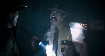 Movie still from “Super Troopers 2” (2018), directed by Jay Chandrasekhar – A man in a uniform and a hat is holding a flashlight; Medium shot, Low angle