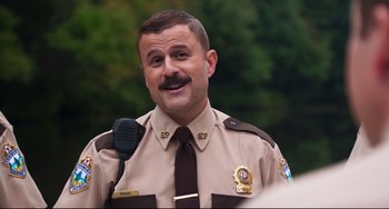 Movie still from “Super Troopers 2” (2018), directed by Jay Chandrasekhar – A man in a brown uniform is holding a microphone and smiling; Close Up shot, Over the shoulder angle