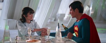 Movie still from “Superman II” (1980), directed by Richard Lester – A man and a woman sitting at a dinner table holding hands; Medium shot, Over the shoulder angle