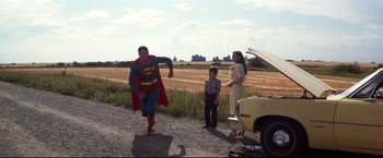 Movie still from “Superman III” (1983), directed by Richard Lester – A man in a superman costume standing next to a woman and a boy; Wide shot, Over the shoulder angle