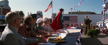Movie still from “Superman III” (1983), directed by Richard Lester – A group of people sitting at a table with flags in the background; Wide shot, Over the shoulder angle