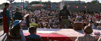Movie still from “Superman III” (1983), directed by Richard Lester – A crowd of people sitting and standing in front of a man in uniform; Wide shot, High angle