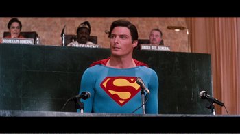 Movie still from “Superman IV: The Quest for Peace” (1987), directed by Sidney J. Furie – A man in a superman costume is standing at a podium in front of other people; Medium shot, Over the shoulder angle