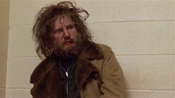Movie still from “Suspect” (1987), directed by Peter Yates – A man with long hair and a beard sitting against a brick wall; Medium shot, Low angle