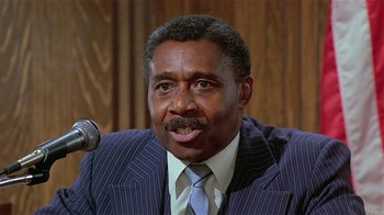 Movie still from “Suspect” (1987), directed by Peter Yates – A man wearing a suit and tie sitting in front of a microphone; Close Up shot, Low angle