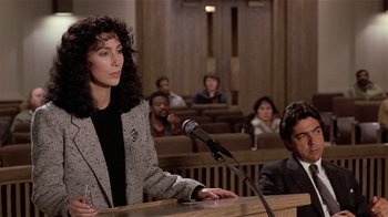 Movie still from “Suspect” (1987), directed by Peter Yates – A woman standing in front of a microphone in a courtroom; Medium shot, Over the shoulder angle