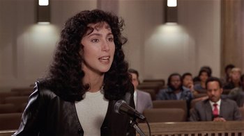 Movie still from “Suspect” (1987), directed by Peter Yates – A woman with curly hair talking into a microphone in front of a group of people; Close Up shot, Over the shoulder angle