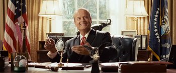 Movie still from “Swing Vote” (2008), directed by Joshua Michael Stern – A man sitting at a table in front of an elephant statue; Medium shot, Low angle