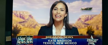 Movie still from “Swing Vote” (2008), directed by Joshua Michael Stern – A woman is smiling for the camera in front of a news desk; Close Up shot, Over the shoulder angle