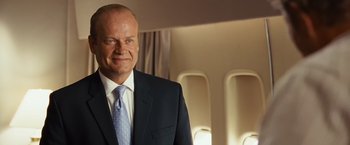 Movie still from “Swing Vote” (2008), directed by Joshua Michael Stern – A man in a suit and tie standing in front of an airplane window; Medium shot, Low angle