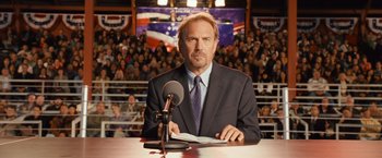 Movie still from “Swing Vote” (2008), directed by Joshua Michael Stern – A man in a suit and tie sitting at a microphone; Medium shot, Low angle