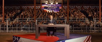Movie still from “Swing Vote” (2008), directed by Joshua Michael Stern – A man sitting at a podium in front of a crowd of onlookers; Wide shot, High angle