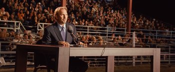 Movie still from “Swing Vote” (2008), directed by Joshua Michael Stern – A man sitting at a podium in front of an audience; Medium shot, Over the shoulder angle